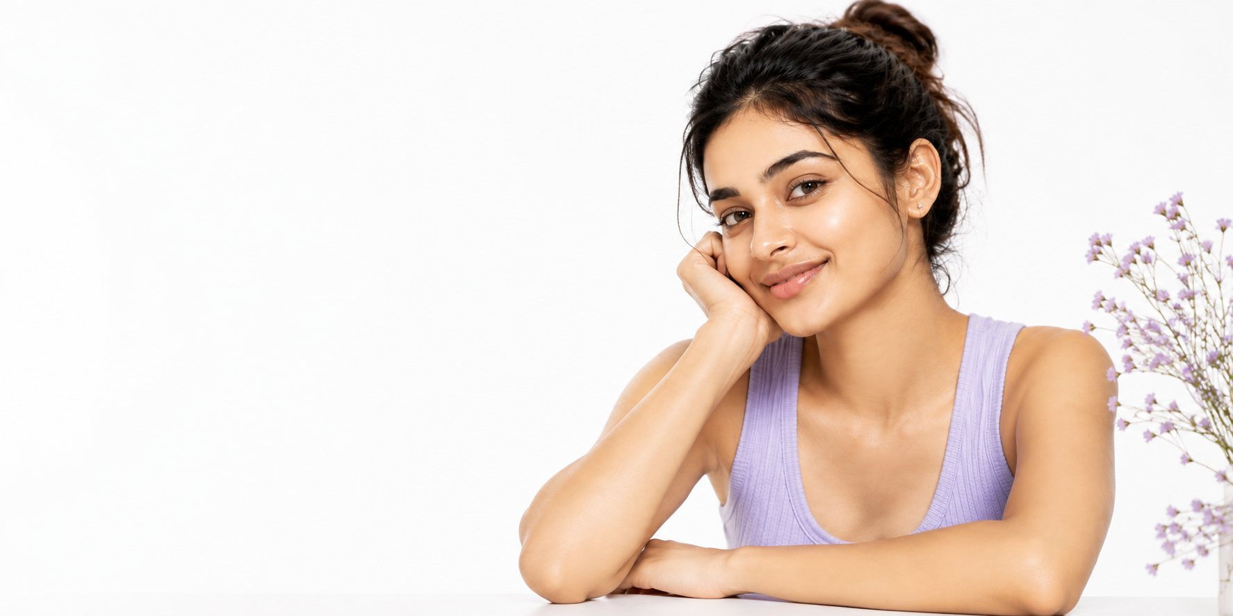 A woman with her hair in a bun wearing a light purple ribbed tank top used baanch skincare, resting her head on her hand and smiling at the camera against a white background with a small floral arrangement on the right.